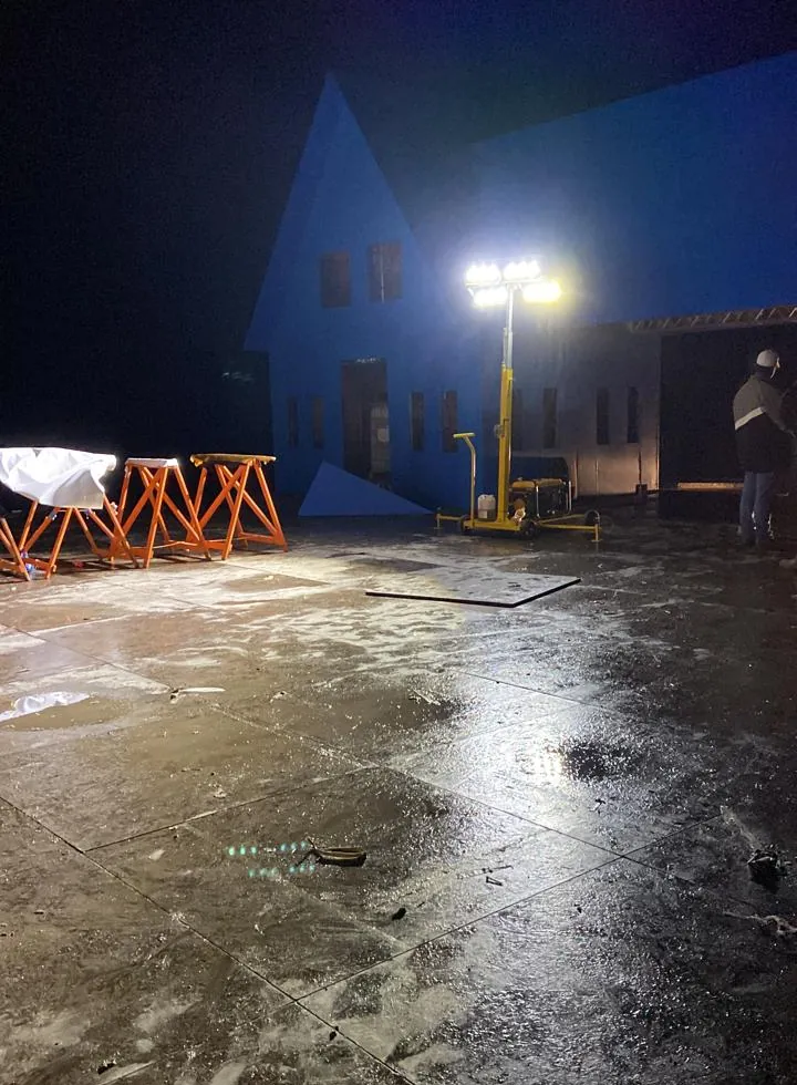 Torre de iluminação Revlo amarela acesa à noite, iluminando um pátio molhado diante de uma estrutura cenográfica azul, em 2022