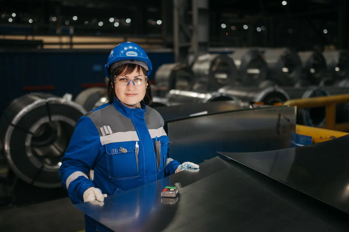 Técnica de uniforme industrial, capacete e óculos de proteção ao lado de bobinas e chapas de aço em galpão